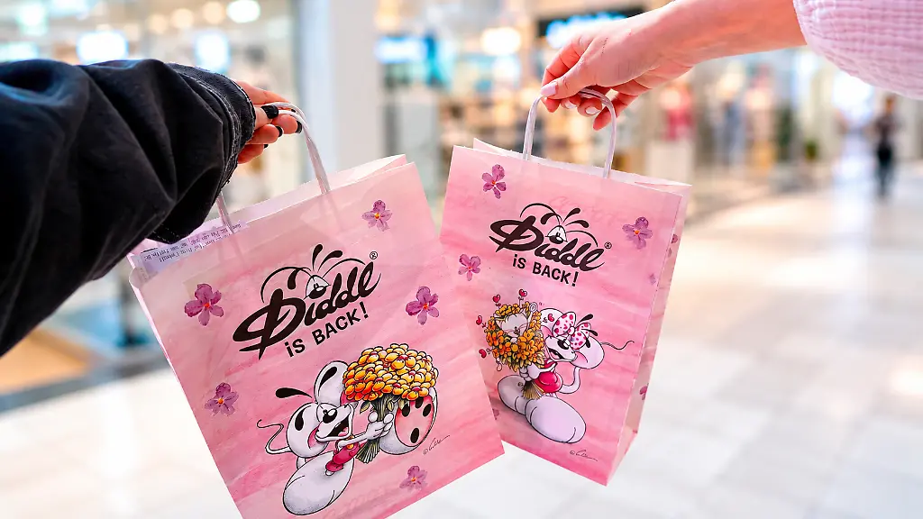 Zwei Hände halten rosafarbene Papiertüten mit dem Aufdruck „Diddl is back!“ in einem Einkaufszentrum.